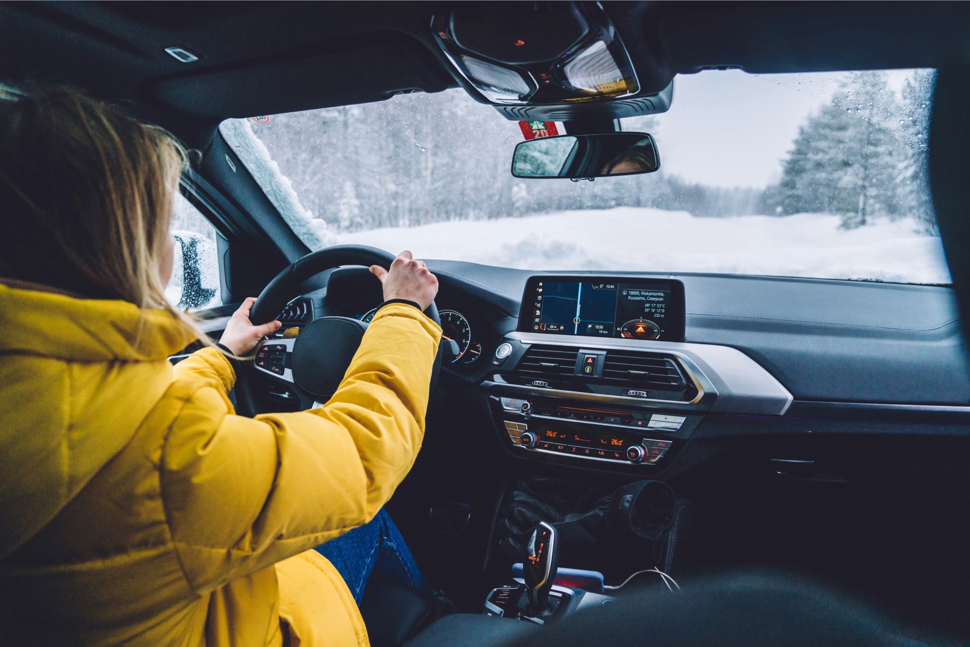 A person wearing a yellow coat drives with two hands on the wheel with a snow filled road beyond the windshield