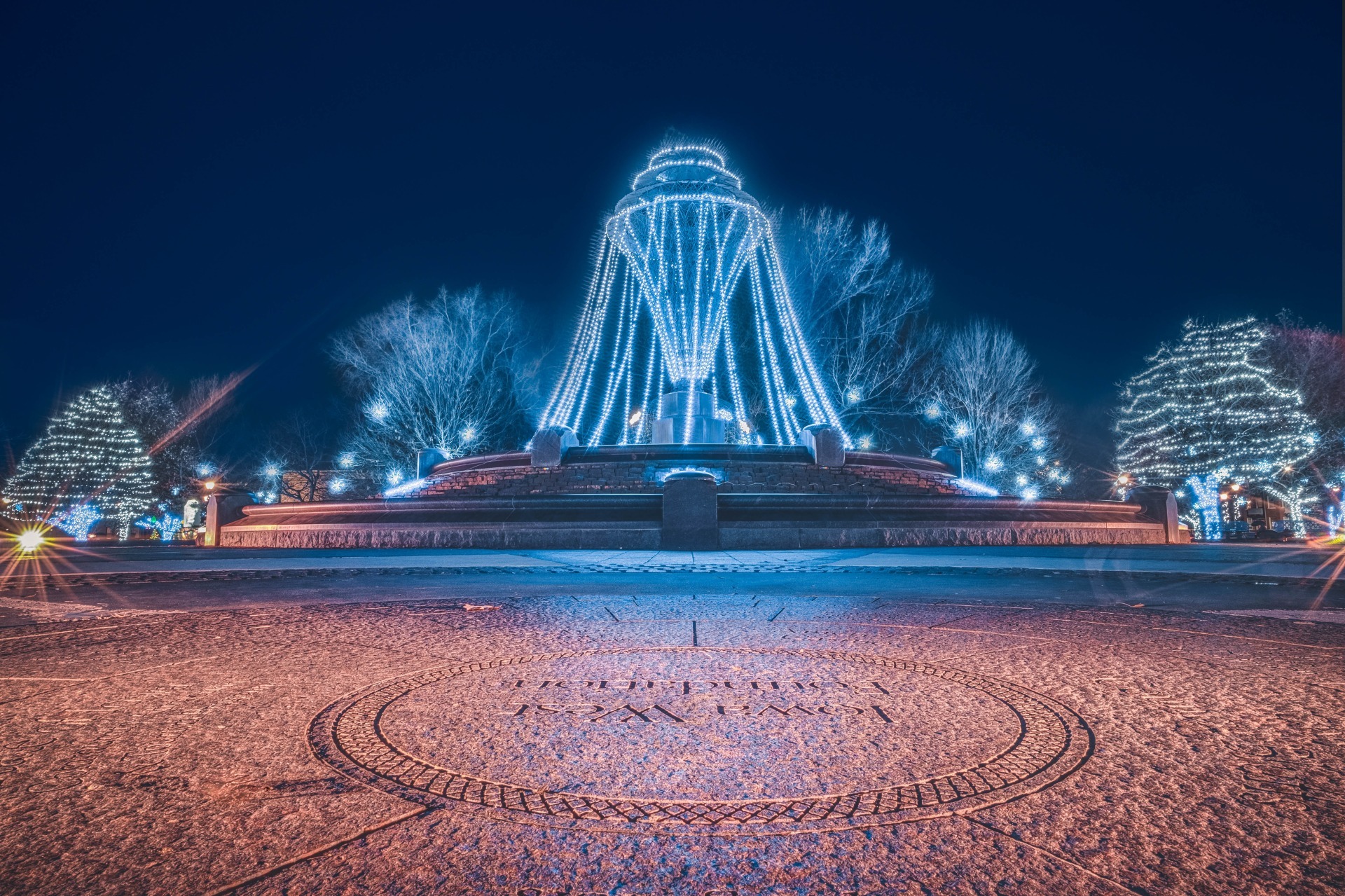 Holiday lights glow a fountain and trees in Bayliss Park