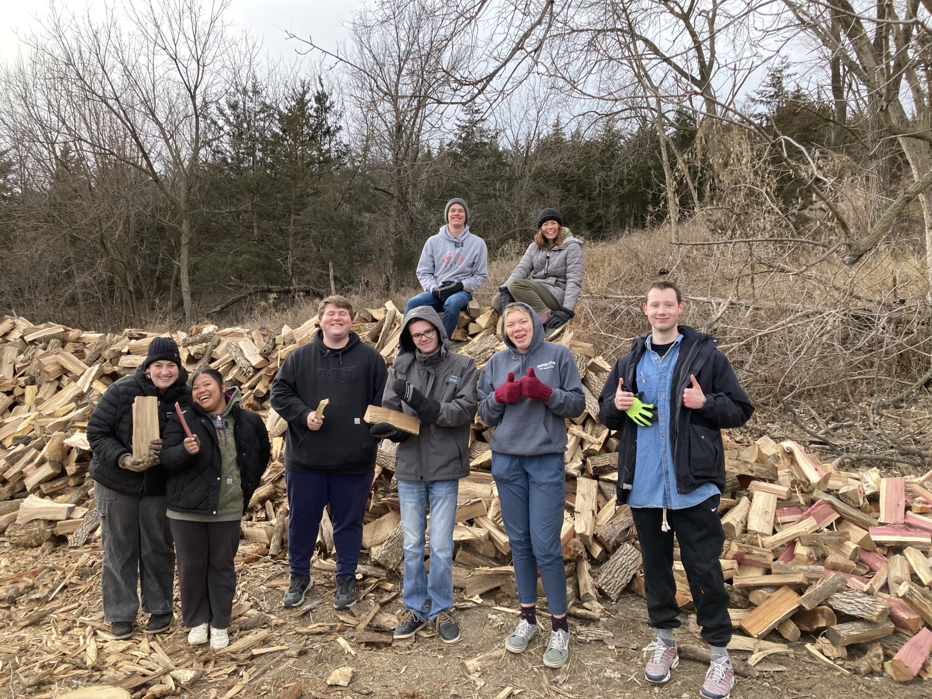 Volunteers assist with firewood splitting at Crescent Hill with large piles of firewood in the background.